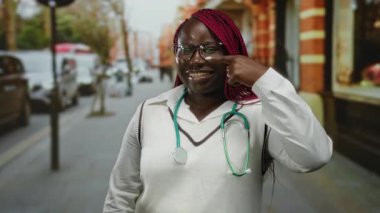 Gözlüklü ve steteskoplu gülümseyen Afro-Amerikan kadın bir şehir caddesinde yanağını gösteriyor, kendine güveni ve sağlık bilincini yansıtıyor..