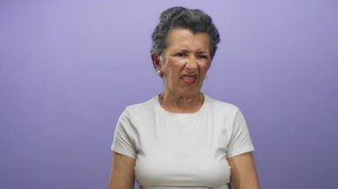 Beyaz tişörtlü yaşlı bir kadın mor arka planda yüzünü buruşturuyor, rahatsızlık ve hoşnutsuzluk anı yakalıyor..
