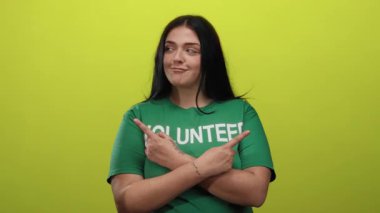 Genç ve cüsseli gönüllü kadın, izole edilmiş sarı arka plan duvarına karşı yeşil gömlek giyerek ve kendinden emin bir ifade sergileyerek her iki tarafı da işaret ediyor..
