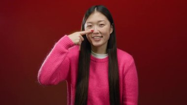 Kırmızı arka planda burnu gösteren pembe kazaklı bir kadın, neşeli bir ifade sergiliyor, izole bir ortamda saçlarıyla Asyalı moda tarzını vurguluyor..