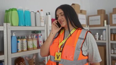 Turuncu yelekli kadın, bağış merkezinde telefonla konuşuyor. Arka planda bir sürü malzeme var..