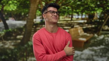 Parkta gülümseyen gözlüklü ve arka planda ağaçlar ve banklar olan kırmızı gömlekli genç bir adam açık havada bir sahne hazırlıyor.