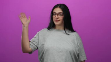 Gözlüklü genç bir kadın canlı pembe bir duvara el sallıyor, kendinden emin bir şekilde gülümsüyor, gri bir gömlek giyiyor, izole bir arka planı var..