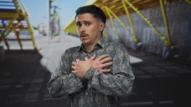 Sokak ortasında oturan genç İspanyol adam. Parlak güneş ışığı altında ve modern şehir geçmişinde elleri göğsünde korkmuş görünüyor..