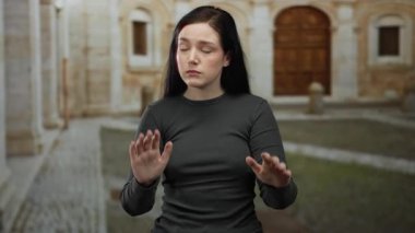 Esmer kadın üniversite kampüsünde etkileyici bir şekilde el kol hareketi yapıyor. Geçmişinde tarihi bir mimari var.