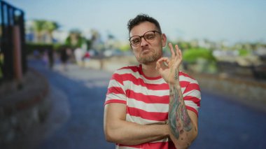 Dövmeli ve gözlüklü genç bir adam, parlak bir şehir sokağında kenar çizgili bir gömlek ve bulanık bir şehir arka planıyla güvenle dururken iyi bir jest yapıyor..