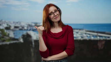 Deniz kenarındaki limanı işaret eden bir kadın, açık havada duruyor, kırmızı giyiyor, açık gökyüzü altındaki deniz ve teknelere karşı kendine güvenen bir ifade sergiliyor..