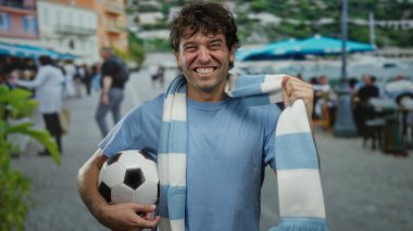 İspanyol adam futbol topu tutuyor ve atkı takıyor. Güneşli bir sokakta heyecanlı bir şekilde gülümsüyor..