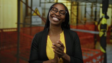 Şantiyede gülümseyen kıvırcık saçlı, gözlüklü, sarı gömlekli ve siyah ceketli bir kadın açık hava endüstriyel ortamda profesyonel güvenini gösteriyor..