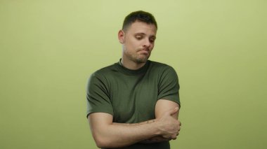 Sakallı ve dövmeli genç adam sarı bir arkaplan karşısında dikiliyor. Düşüncesizce hareket ediyor ve yeşil bir gömlek giyiyor..