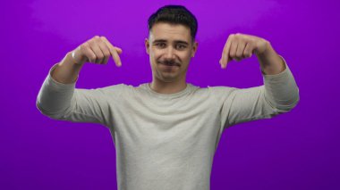 Genç İspanyol adam iki parmağıyla aşağıya doğru işaret ediyor. Kalın mor arka plan duvarına karşı. Açık gri gömlek giyiyor.