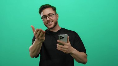 Sakallı ve gözlüklü genç adam tecrit edilmiş yeşil duvar arkaplanına karşı akıllı telefon kullanıyor. Teknoloji ve iletişim üzerine odaklanmış çeşitli ifadeler sergiliyor..