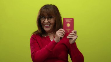 Kıdemli İspanyol kadın, elinde canlı sarı bir arka plana karşı Alman pasaportu tutarken gülümsüyor. Seyahat ve kimlik temalarını örnek alıyor..