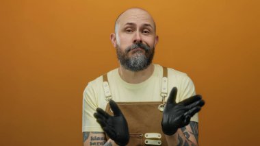 Turuncu arka planda kahverengi önlüklü sakallı bir adam ciddi bir ifade sergiliyor..