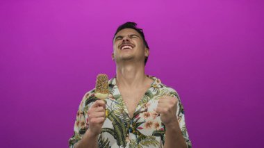 İspanyol asıllı genç adam pembe arka planda, çiçekli gömlekli ve modern tarzda neşeli yaz kaçışını yansıtan pembe elbisesiyle dondurma keyfine varıyor.