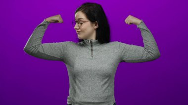 Gözlüklü, kendine güvenen, yumruklarını kaldırmış, mor bir arka plana karşı poz veren, gri bir süveter giyen, güç, güç ve kararlılık gösteren bir kadın..