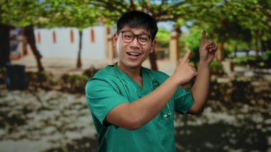 Önlüklü, steteskoplu genç Çinli adam gülüyor ve arka planda ağaçlar ve gölgeler olan güneşli bir parkı işaret ediyor..