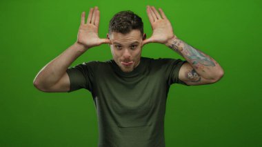 Alaycı gülüşü ve rahat duruşu olan yeşil bir arka planda oyuncu ifadeler sergileyen genç bir adam.