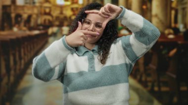 Gözlüklü genç İspanyol kadın kilisenin içinde ahşap banklar ve arka planda sıcak ışıklandırmayla bir çerçeve hareketi yapıyor..