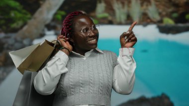 Açık havuz tesisini işaret eden alışveriş çantalarıyla gülümseyen kadın, arka planda açık mavi su bulunan canlı tatil atmosferinde keyif ve keyfi sembolize ediyor..