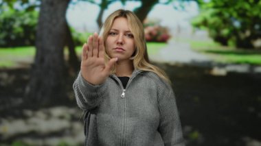 Parkta ciddi bir ifadeyle duran, el kaldıran, sarı saçlı ve gri kazaklı bir kadın..