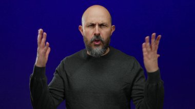 Kel sakallı bir adam, mavi arka plana karşı öfkesini dışa vuruyor, yüz ifadesi ve hareketlerinde duygu ve yoğunluk ifade ediyor..