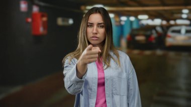 Loş bir otoparkta kapalı bir yeri işaret eden kadın, günlük kıyafetler giyiyor, gizemli bir ambiyansı olan arabaların yanında meraklı ve meraklı bir ifade sergiliyor..
