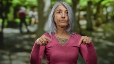 Gri saçlı, pembe gömlekli bir kadın açık hava parkı ortamında düşünceli bir şekilde jestler yapıyor. Canlı yeşillik ve yumuşak ışığın arasında bilgelik ve karakter aktarıyor..