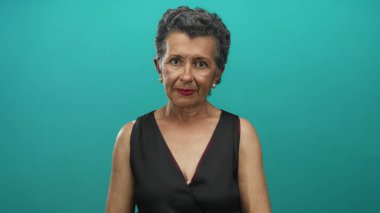 Gri saçlı, siyah elbiseli yaşlı bir kadın mavi arka planda kameraya bakıyor. Stüdyo ortamında güven ve haysiyet taşıyor.