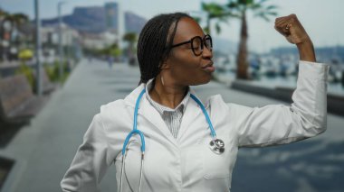 Beyaz önlüklü ve gözlüklü Afrikalı Amerikalı kadın doktor, deniz kenarındaki gezinti güvertesinde steteskopla güvenle duruyor..