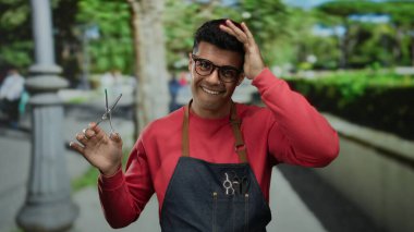 Genç İspanyol asıllı bir adam elinde makasla, kırmızı bir kazak ve önlükle dışarıda poz veriyor. Şehir arka planı hareketli ve davetkar bir atmosferi arttırıyor..