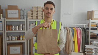 Yansıtıcı yelek giyen genç bir gönüllü, içinde yiyecek ve giyecek dolu raflarla çevrili hayır kurumu bağış merkezinde kağıt torba tutuyor.