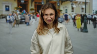 Şaşkın bakışlı genç bir kadın kalabalık bir şehir caddesinde duruyor. Etrafı kalabalıkla çevrili, beyaz bir süveter giyiyor. Gün ortasında açık havada..