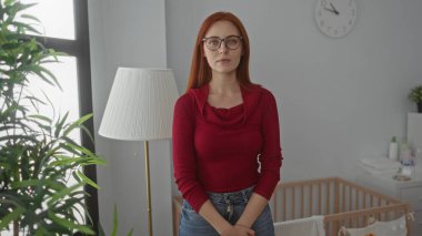 Kırmızı kazaklı bir kadın, yatak odasında rahat bir yatak odasında duruyor. Kapalı bitkiler ve beyaz lamba, ev kreşinin huzurlu atmosferini yansıtıyor..