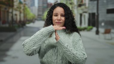 Şehir caddesinde yeşil süveter giyen bir kadın, duraklama sinyali veriyor ya da gündüz vakti şehir ortamında odaklanmış bir ifadeyle duruyor..