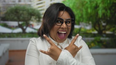 Latin kökenli bir kadın, arka planda canlı yeşillik olan bir parkta neşeyle kaya jesti yapıyor, gözlük takıyor ve beyaz bir gömlek giyiyor, enerjik bir şekilde gülümsüyor..