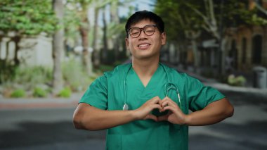 Yeşil önlüklü genç Çinli bir adam bir şehir caddesinde kalp jesti yapıyor, boynunda steteskopla dışarıda gülümsüyor, neşeli ve dost canlısı bir sağlık çalışanını somutlaştırıyor..