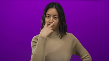 Genç bir kadın, canlı mor bir arkaplan karşısında iki parmağıyla ihtiyatlı bir jest yapar. Farkındalık ve odaklanma duygusunu aktarır..