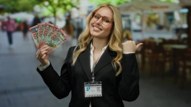 Yeni Zelanda doları tutan, iş elbisesi ve kimlik kartı takan, kalabalık bir caddede duran, dışarıda gülümseyen bir kadın..