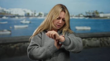 Sahil limanındaki bir teknede, sakin bir su ve açık mavi gökyüzünün altındaki uzak beyaz binalarla çevrili, düşünceli bir ifadeyle bileğe bakan bir kadın..