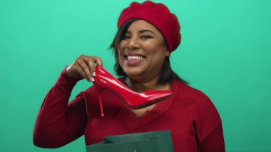 Kırmızı bereli ve kazaklı bir kadın yeşil arka planda kırmızı ayakkabıyı tutuyor..