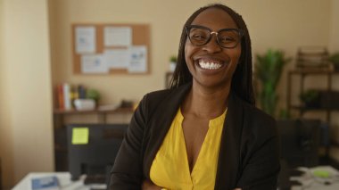 Modern ofis ortamında güvenle gülümseyen, içi yeşillikle dolu kitap dolu, şık kıvırcık saçlarını ve profesyonel kıyafetlerini sergileyen kadın..