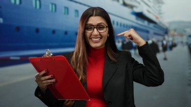 Limanda pano ile gülümseyen, gözlüklü, arka planda insanlarla birlikte büyük teknenin yanında duran, canlı bir ortamda kendine güvenen ve profesyonellik sergileyen bir kadın..