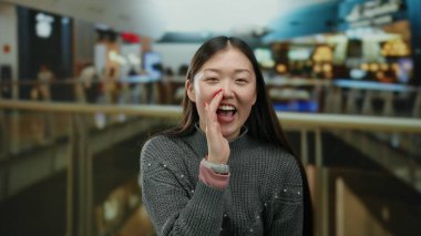 Arka planda canlı mağazalar olan hareketli bir alışveriş merkezinde bağıran, rahat bir kazak giyerken heyecan ve neşe ifade eden bir kadın..