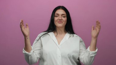 Büyük beden bir kadın, beyaz gömlekli, elleri pembe arka planda, kendine güveni ve zarafeti canlı bir ortamda somutlaştırıyor..