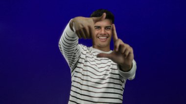 Genç adam çizgili bir gömlek içinde canlı mavi bir arka plana gülümsüyor, elleriyle çerçeveletiyor, oyuncu bir duruş sergiliyor ve çekici, kendinden emin bir tavır sergiliyor..