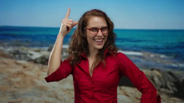 Kırmızı gömlekli kadın deniz kenarında neşeyle gülüyor, açık mavi deniz arka planına işaret ediyor, kumsaldaki mutluluk ve heyecanı somutlaştırıyor..