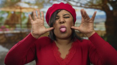 Kırmızı bereli genç ve iri yarı Latin bir kadın gündüz vakti parkta komik suratlar yapıyor..