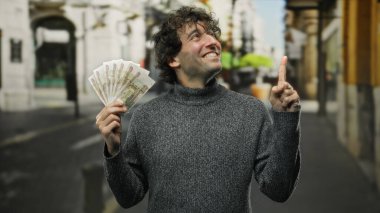 İspanyol adam elinde Rus rublesi tutarken gülümsüyor ve canlı bir şehir caddesini işaret ediyor. Açık havada iyimser ve finansal bir başarı havası yakalıyor..