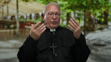 Kıdemli rahip gözlüklü bir adam sokakta jest yapıyor. Arka planda bulanık yeşil ağaçlar ve insanlar var. Huzurlu bir ruhaniyet hissi taşıyor..